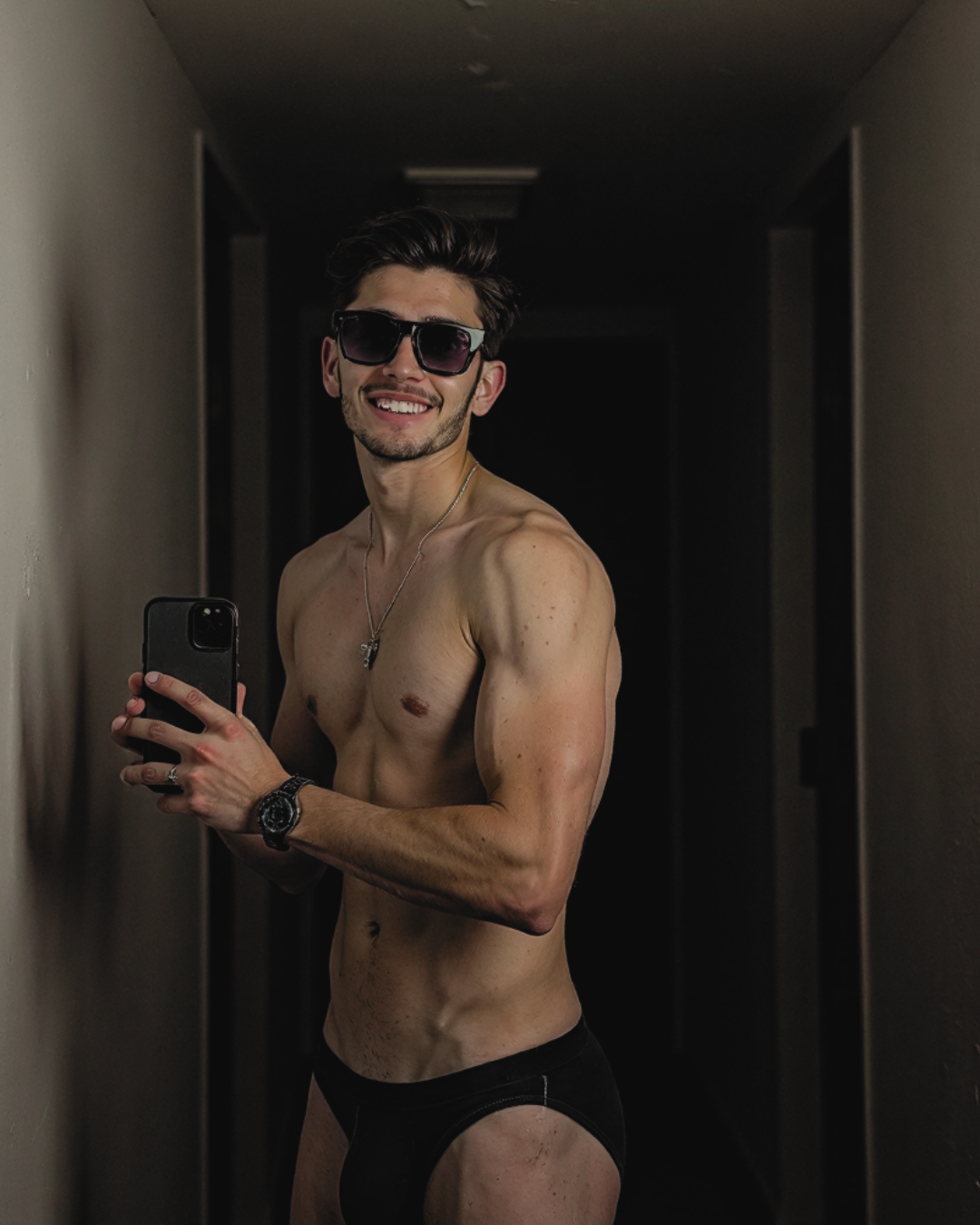 Man taking a mirror selfie wearing sunglasses and black underwear in a dimly lit room.