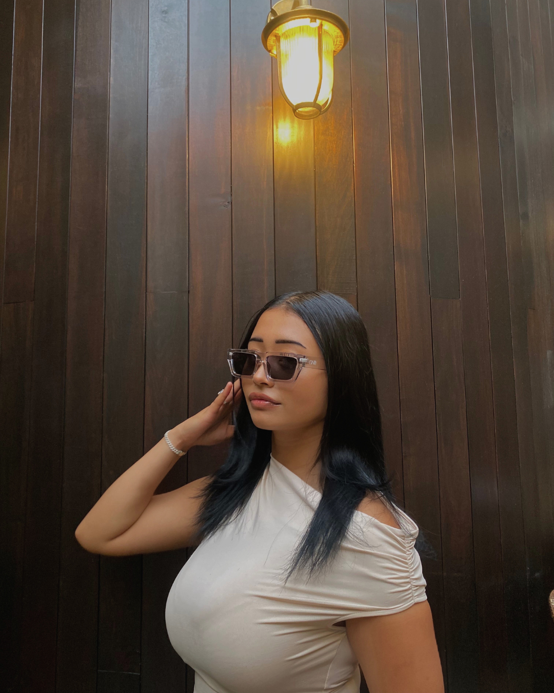 Woman wearing sunglasses against a wooden wall with a lamp.